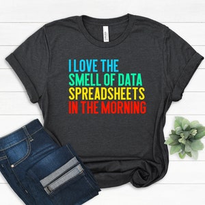May include: Dark gray t-shirt with the text "I LOVE THE SMELL OF DATA SPREADSHEETS IN THE MORNING" in blue, green, yellow, and red. The shirt is paired with blue jeans.