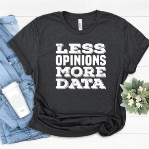 May include: Dark gray t-shirt with the text "LESS OPINIONS MORE DATA" in white block letters. The shirt is folded, with a denim jacket and a white coffee cup visible in the background. A small plant is also in the background.