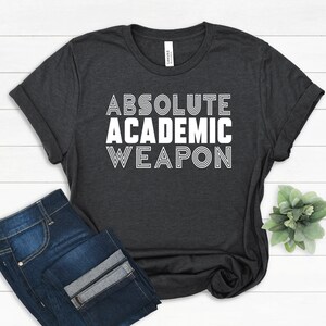 Może przedstawiać: Ciemnoszara koszulka z białym napisem "ABSOLUTE ACADEMIC WEAPON". Koszulka jest połączona z niebieskimi dżinsami. Rękawy koszulki są podwinięte.
