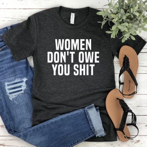 Pode incluir: T-shirt cinza escuro com o texto branco "WOMEN DON'T OWE YOU SHIT". Combinado com jeans azuis e sandálias pretas. Uma pequena planta verde é visível no fundo.