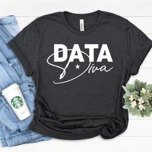 Könnte beinhalten: Dunkelgraues T-Shirt mit dem weißen Schriftzug "DATA Diva" und einem Stern. Die Ärmel sind umgeschlagen. Eine Jeansjacke und ein Starbucks-Becher liegen neben dem T-Shirt.