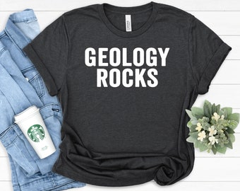 Puede incluir: Una camiseta gris oscuro con el texto "GEOLOGY ROCKS" impreso en blanco.