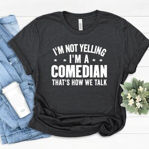 May include: Dark grey t-shirt with the white text "I'M NOT YELLING I'M A COMEDIAN THAT'S HOW WE TALK". The shirt is folded, with a denim jacket and a white coffee cup visible in the background. A small plant is also in the background.