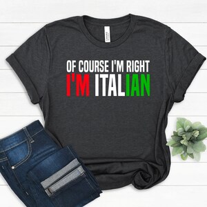 May include: Dark gray t-shirt with the text "OF COURSE I'M RIGHT I'M ITALIAN" in white, red, and green. The shirt is folded, with a pair of blue jeans visible in the lower left corner. A small plant is in the lower right corner.