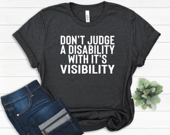 Puede incluir: Camiseta gris oscuro con el texto blanco "DON'T JUDGE A DISABILITY WITH IT'S VISIBILITY". La camiseta se combina con vaqueros azules. La camiseta tiene mangas enrolladas y cuello redondo.