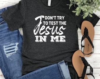 May include: A dark charcoal heather gray short-sleeve t-shirt featuring the white printed text "DON'T TRY TO TEST THE JESUS IN ME" in a mix of bold block and script lettering, paired with blue denim jeans and black braided strap sandals.