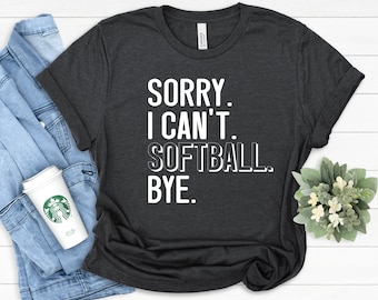 Op de afbeelding: Een donker antracietgrijs t-shirt met korte mouwen en de witte opdruk SORRY. I CAN'T. SOFTBALL. BYE. Het t-shirt wordt getoond naast een blauw spijkerjack, een witte koffiebeker met een groen logo en een kleine groene plant.