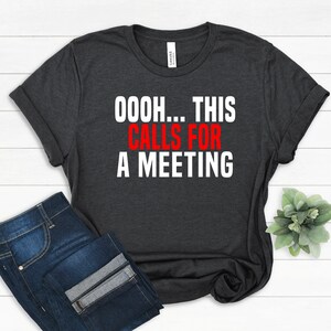 May include: A dark charcoal grey short-sleeve t-shirt with white and red text printed on the front that reads, "OOOH... THIS CALLS FOR A MEETING." The shirt features a crew neckline and a standard fit, displayed flat with folded sleeves.