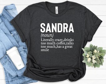 Può includere: T-shirt grigio scuro con il nome "SANDRA" in lettere bianche, seguito da una definizione. La definizione recita: "Letteralmente pazza, beve troppo caffè, parla troppo, ha un grande sorriso."