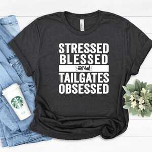 May include: Dark gray t-shirt with the text "STRESSED BLESSED and TAILGATES OBSESSED" in white. The shirt is folded, with a denim jacket and a coffee cup visible in the background. The shirt is made of a soft material.
