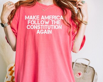 Puede incluir: Una camiseta de color coral con el texto "MAKE AMERICA FOLLOW THE CONSTITUTION AGAIN" en letras blancas. La camiseta tiene mangas cortas y un corte relajado, adecuada para uso diario.