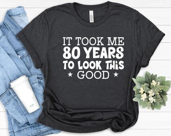 Puede incluir: Una camiseta de manga corta en color gris carbón oscuro con el texto impreso en blanco "IT TOOK ME 80 YEARS TO LOOK THIS GOOD" y una estrella a cada lado de la palabra "GOOD", centrado en la parte frontal con una tipografía redondeada.