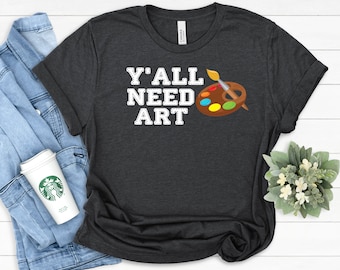 May include: A dark charcoal gray short-sleeve t-shirt featuring the white text Y'ALL NEED ART in a stitched font style. To the right of the text is a brown wooden artist palette with colorful paint dabs and a paintbrush resting on top.