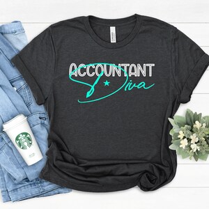 Könnte beinhalten: Ein dunkelgraues T-Shirt mit dem Text "Accountant Diva" in türkisblauer Schrift. Der Text ist in einer stilisierten Schriftart mit einem Stern und einer Stiftgrafik.