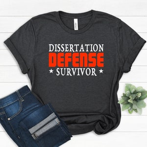 May include: Dark gray t-shirt with the words "DISSERTATION DEFENSE SURVIVOR" in white and red block letters. The shirt is folded, with a pair of blue jeans visible below. A small plant is in the background.