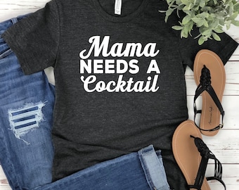 Puede incluir: Camiseta gris oscuro con la frase "Mama Needs A Cocktail" en escritura blanca. La camiseta se combina con vaqueros azules y sandalias negras. Las sandalias tienen un diseño de correa trenzada. La imagen está tomada sobre una superficie de madera blanca.