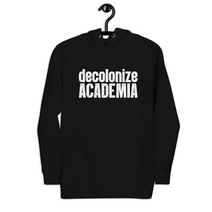 Pode incluir: Uma camisola com capuz preta com o texto branco "decolonize ACADEMIA" impresso no peito. A camisola inclui um capuz com cordão, mangas compridas e um bolso frontal tipo canguru num design padrão.