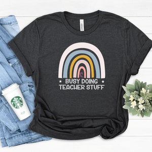 Puede incluir: Una camiseta gris marengo jaspeado con un gráfico de un arcoíris multicolor en tonos rosa, azul y amarillo. Debajo del arcoíris, el texto "BUSY DOING TEACHER STUFF" aparece en letras mayúsculas blancas, flanqueado por dos pequeñas estrellas.