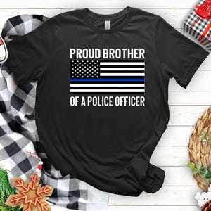 Puede incluir: Camiseta negra con una bandera estadounidense blanca con una franja azul en el medio. El texto "Proud Brother of a Police Officer" está impreso debajo de la bandera.
