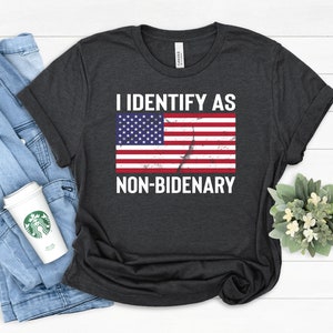 Puede incluir: Una camiseta de manga corta de color gris carbón oscuro con el texto blanco I IDENTIFY AS NON-BIDENARY encima y debajo de un gráfico desgastado de la bandera de los Estados Unidos. La camiseta tiene cuello redondo y corte estándar.