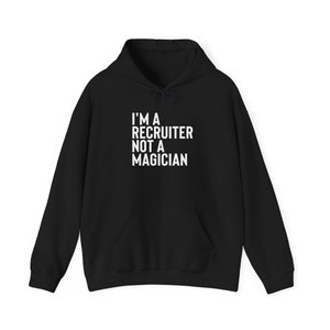 Peut inclure: Sweat à capuche noir avec une poche avant et un cordon de serrage. Le texte blanc sur le devant indique "I'M A RECRUITER NOT A MAGICIAN". Un vêtement décontracté et confortable pour un usage quotidien.