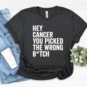 Puede incluir: Camiseta gris oscuro con el texto blanco "HEY CANCER YOU PICKED THE WRONG B*TCH". La camiseta se muestra sobre una superficie blanca con una chaqueta vaquera y una taza de café blanca.
