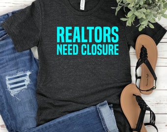 May include: A dark heather gray short-sleeve t-shirt with the text REALTORS NEED CLOSURE printed in bright cyan blue. The shirt is paired with blue denim jeans and a pair of black braided strap sandals, creating a casual ensemble for daily wear.