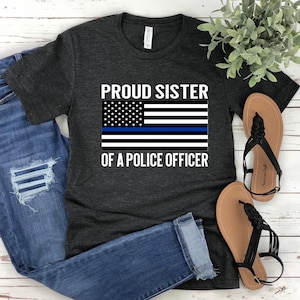 May include: A dark grey t-shirt with the text "Proud Sister of a Police Officer" and an American flag with a blue stripe across it.