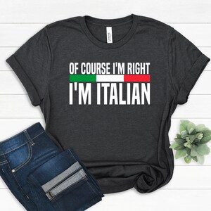May include: Dark gray t-shirt with the text "OF COURSE I'M RIGHT I'M ITALIAN" in white, with the Italian flag colors above the text. The shirt is paired with blue jeans.