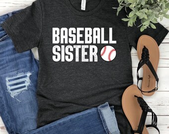 May include: A dark charcoal gray short-sleeve t-shirt featuring the white text BASEBALL SISTER with a graphic of a white baseball with red stitching. The shirt is paired with blue denim jeans and a pair of black braided strap sandals.
