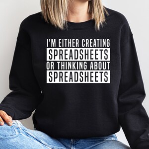May include: Black sweatshirt with white text: "I'M EITHER CREATING SPREADSHEETS OR THINKING ABOUT SPREADSHEETS." The text is in a bold, sans-serif font. The sweatshirt is a solid black colour.