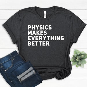 Puede incluir: Camiseta gris oscuro con mangas enrolladas y el texto "PHYSICS MAKES EVERYTHING BETTER" en blanco. La camiseta se combina con unos vaqueros azules. Una pequeña planta verde es visible en el fondo.