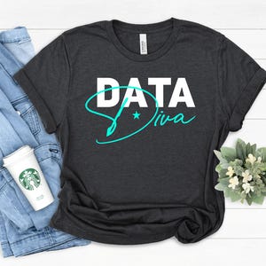 Könnte beinhalten: Dunkelgraues T-Shirt mit dem Schriftzug "DATA Diva" in Weiß und Türkis. Das Shirt ist gefaltet, mit einer Jeansjacke und einer Kaffeetasse im Hintergrund. Eine kleine Pflanze befindet sich in der unteren rechten Ecke.