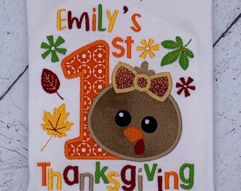 May include: White t-shirt with colorful embroidered text and graphics. The design includes the text "Emily's 1st Thanksgiving" with a turkey face, a number 1, and fall leaves. The shirt is a festive design for a first birthday.
