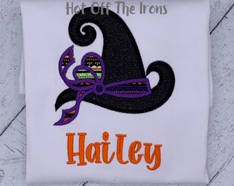 May include: A white t-shirt featuring an embroidered black witch hat with a purple and patterned ribbon. Below the hat, the name Hailey is embroidered in orange thread. The text Hot Off The Irons is printed in gray above the witch hat design.