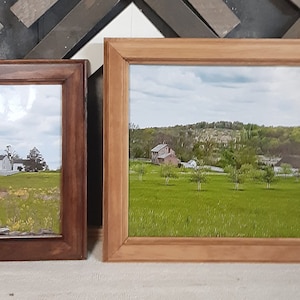 May include: Two framed pictures, one with a brown frame and the other with a light wood frame. The picture in the brown frame shows a white farmhouse in a field with a cloudy sky. The picture in the light wood frame shows a green field with trees and a house in the distance.