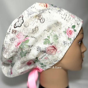 May include: A white surgical scrub cap with a vintage floral and butterfly print. The cap features pink roses, green leaves, and a butterfly design. The cap has a pink ribbon tie and the text "Gypsy Fire & Lace".
