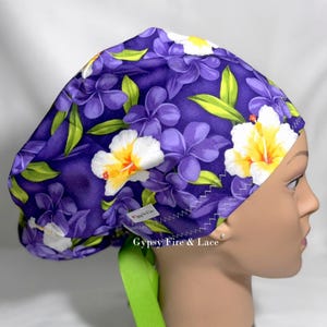 May include: A purple surgical scrub cap with a floral pattern of white and yellow hibiscus flowers and green leaves. The cap has a green tie and a white label with the text "Gypsy Fire & Lace".