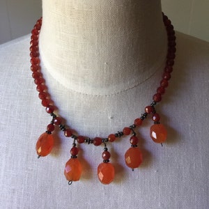 May include: A red beaded necklace with faceted orange pendant beads. The necklace is displayed on a white mannequin. The necklace has a delicate design with small beads and larger, teardrop-shaped pendants.