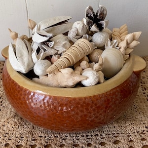 May include: A decorative bowl filled with dried potpourri. The bowl is a textured, reddish-brown color with a light tan rim. The potpourri includes various natural elements in shades of beige and brown, such as seed pods, pine cones, and wooden spirals.