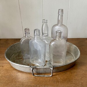 May include: Five clear glass bottles of various shapes and sizes are arranged on a silver metal tray with a handle. The tray is on a wooden surface.