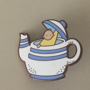 May include: A whimsical enamel pin featuring a baby inside a teapot. The teapot is white with blue stripes and a blue lid. The baby is wearing a yellow outfit. The pin has a copper-colored border.