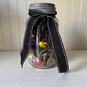 May include: A clear glass jar filled with small trinkets, including a yellow bee charm, a red heart, and a metal initial. The jar is topped with a metal lid and tied with a black ribbon with white stitching.