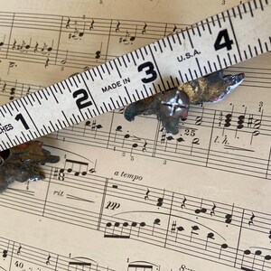 May include: Two small, weathered metal leaf-shaped decorative fasteners with screw-in backs. A white measuring tape is positioned across the items, showing each piece measures approximately 1.5 inches in width from tip to tip.
