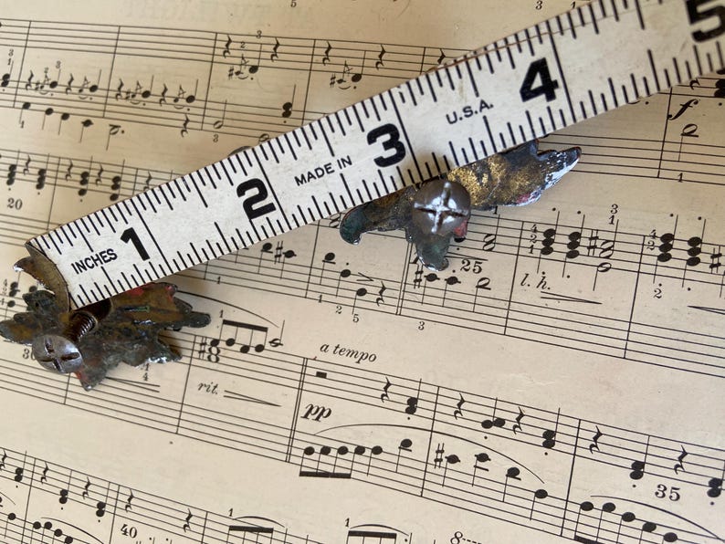 May include: Two small, weathered metal leaf-shaped decorative fasteners with screw-in backs. A white measuring tape is positioned across the items, showing each piece measures approximately 1.5 inches in width from tip to tip.