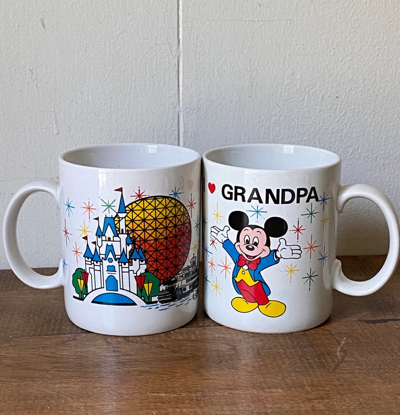 Puede incluir: Dos tazas de cer&aacute;mica blanca con dise&ntilde;os coloridos. Una taza presenta una representaci&oacute;n del castillo de Cenicienta y el paseo Spaceship Earth en Walt Disney World. La otra taza presenta a Mickey Mouse y el texto "Te quiero abuelo".