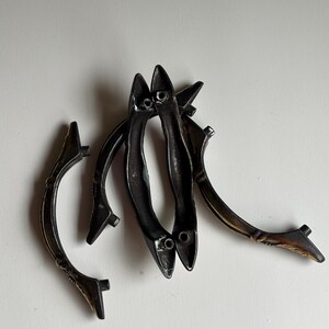 May include: A collection of four curved metal drawer pulls with a dark, weathered finish. Each handle features a slender, arched design with two mounting points at the ends. The metal has a textured, aged appearance with subtle bronze highlights.