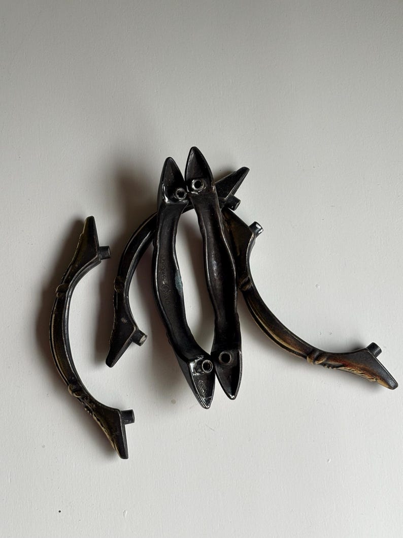 May include: A collection of four curved metal drawer pulls with a dark, weathered finish. Each handle features a slender, arched design with two mounting points at the ends. The metal has a textured, aged appearance with subtle bronze highlights.