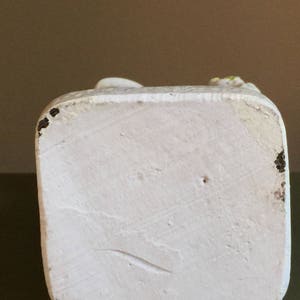 May include: White ceramic figurine with a smooth, textured surface. The figurine is a standing figure with a raised arm and hand. The base of the figurine is square and has a slightly rough texture.