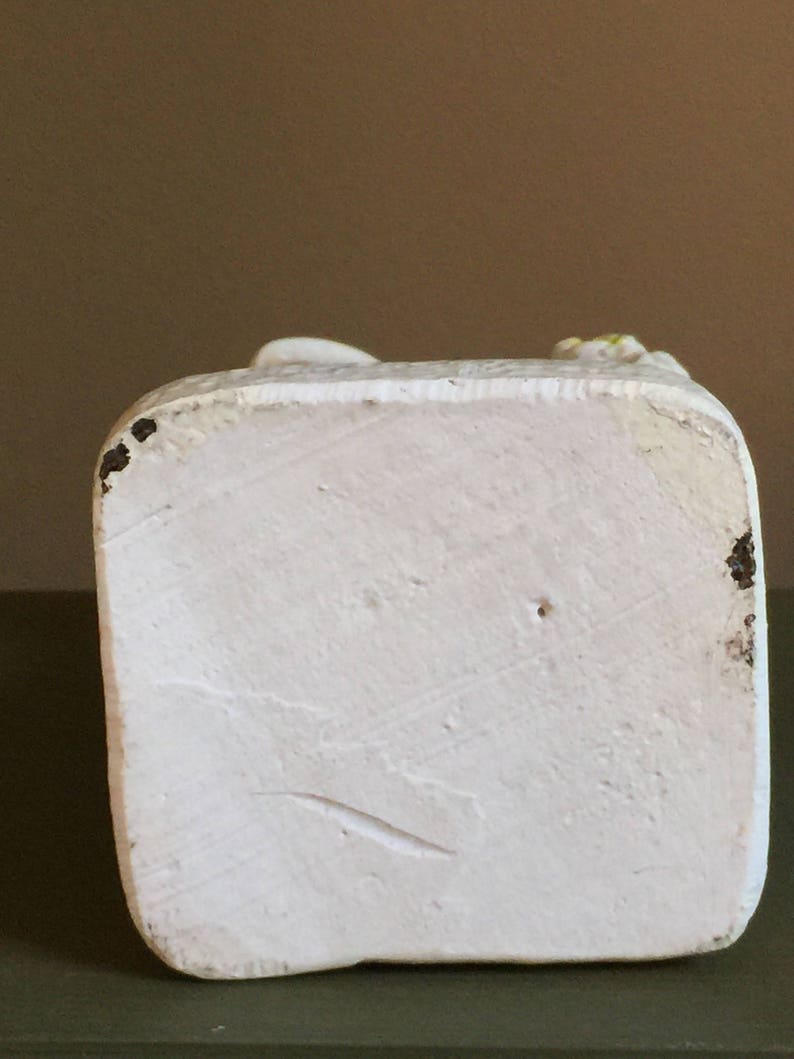 May include: White ceramic figurine with a smooth, textured surface. The figurine is a standing figure with a raised arm and hand. The base of the figurine is square and has a slightly rough texture.